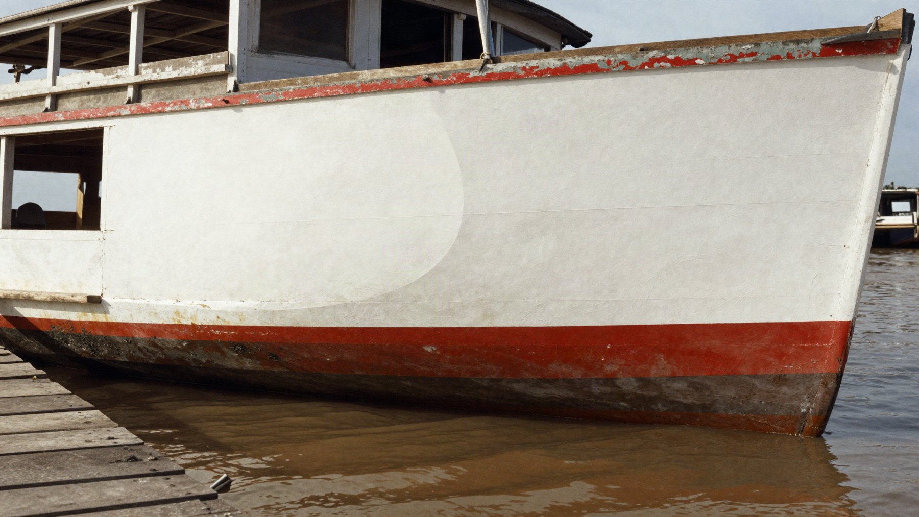 Casco de barco ribeirinho pintado de branco com faixa vermelha, encostado no trapiche sobre o rio barrento